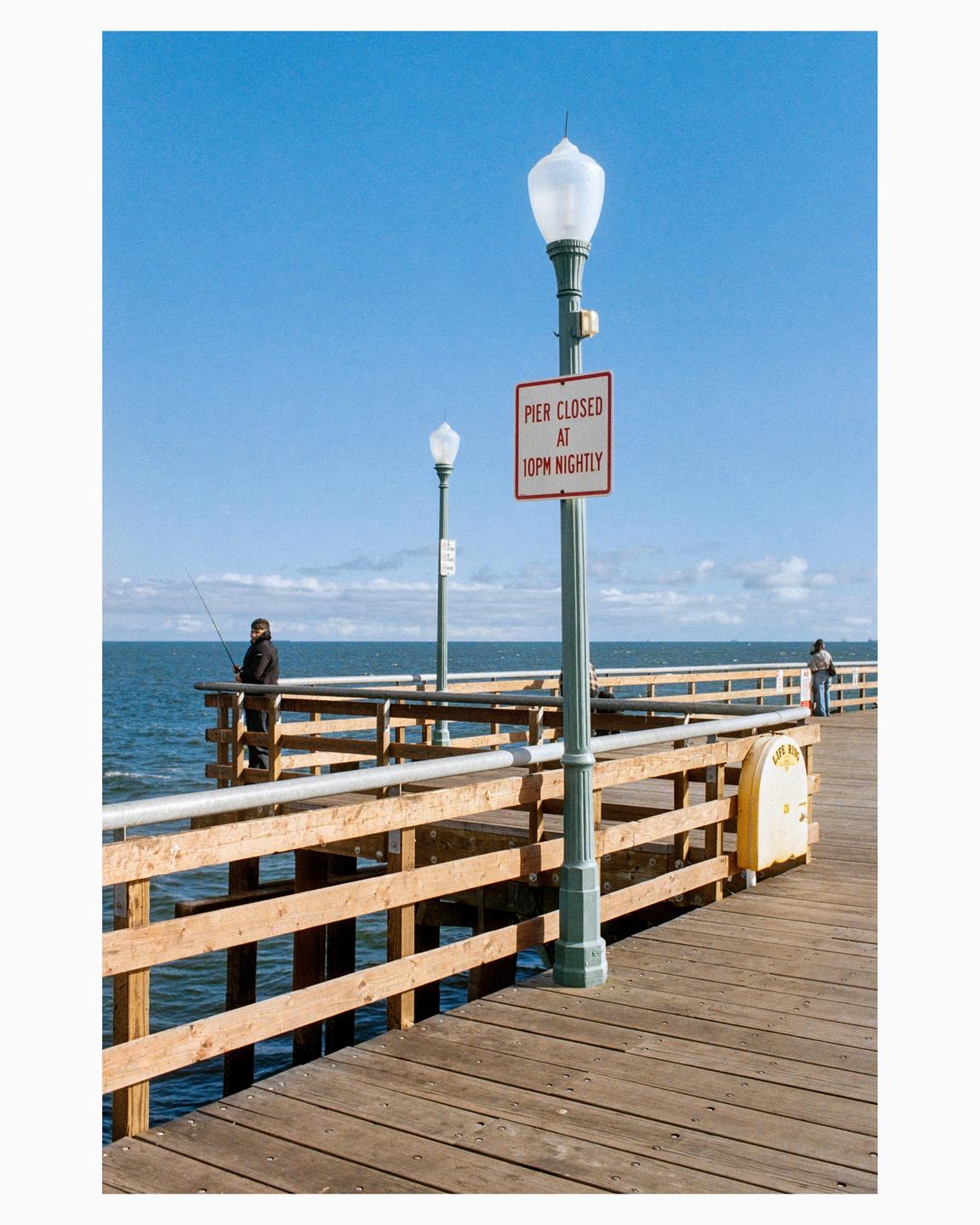 A man fishes from a wooden pier under a clear blue sky with a "pier closed at 10pm nightly" sign visible.