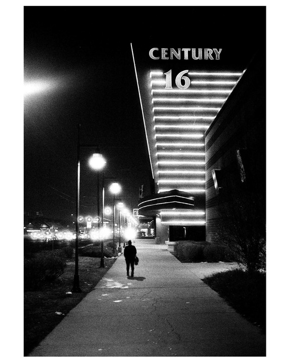 A person walks along a sidewalk at night towards a building with a "Century 16" sign.
