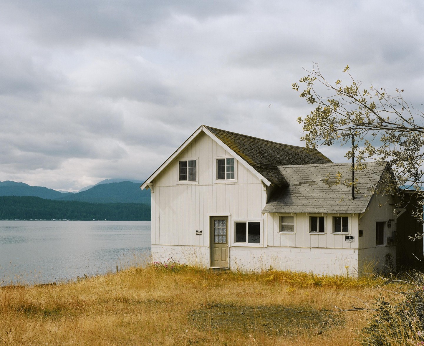 A white house with a mossy roof stands on a grassy hill overlooking a body of water with forested mountains in the distance under a cloudy sky.