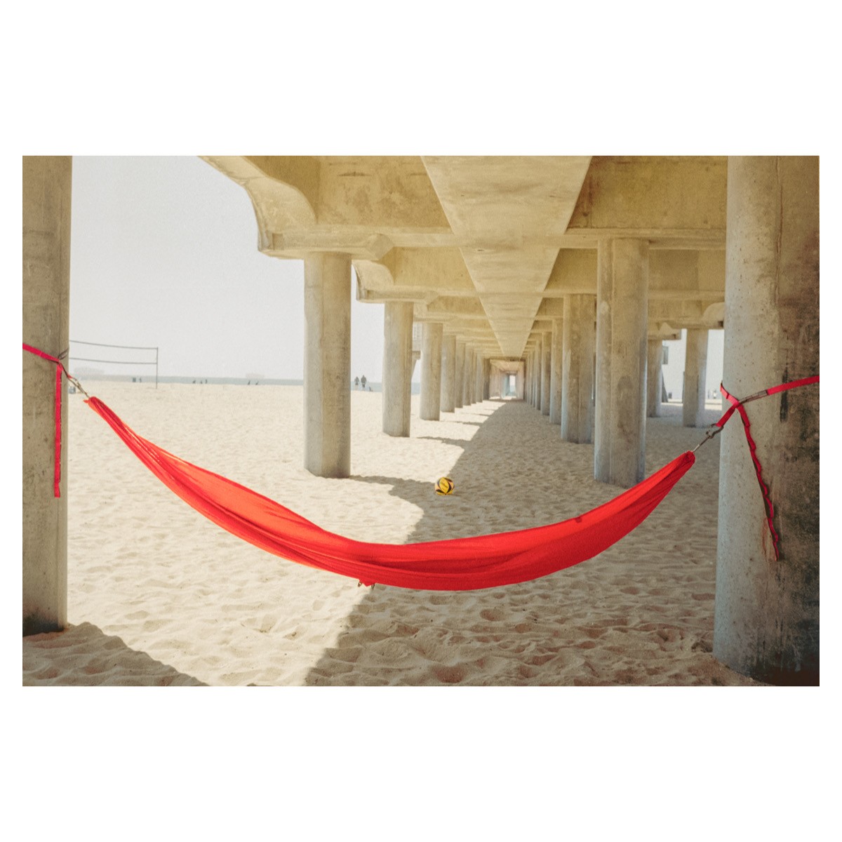 A red hammock is strung between two concrete pillars under a pier on a sandy beach.