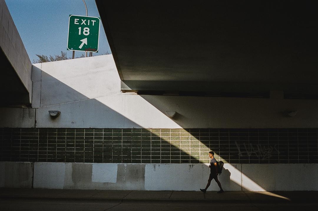 A man walks along a sidewalk under an overpass with a bright exit sign above.