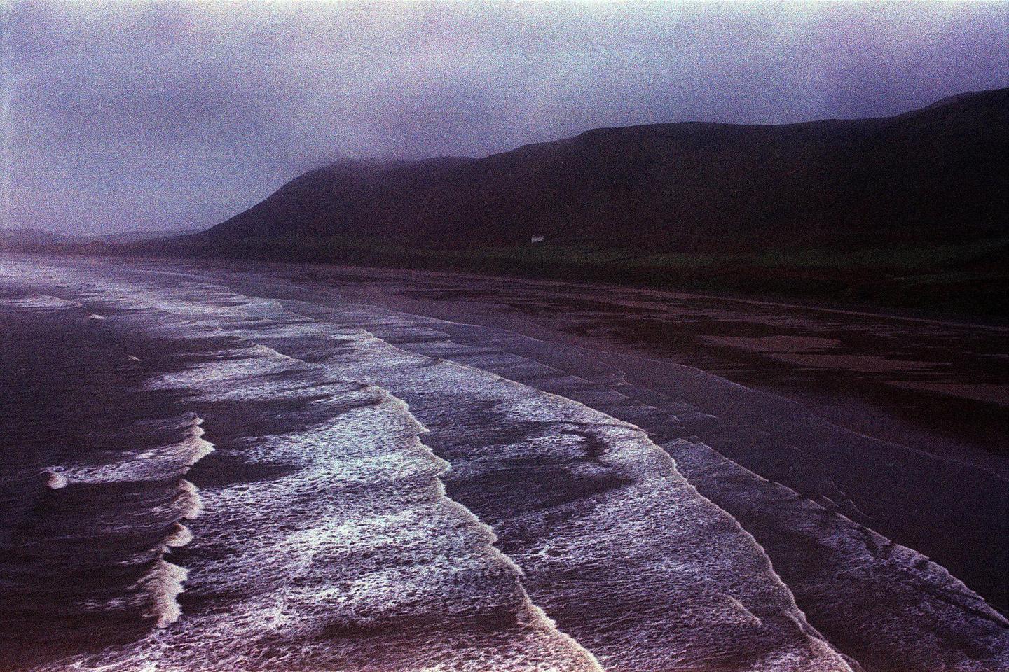 Waves crash on a beach with a mountain in the background under a hazy sky.
