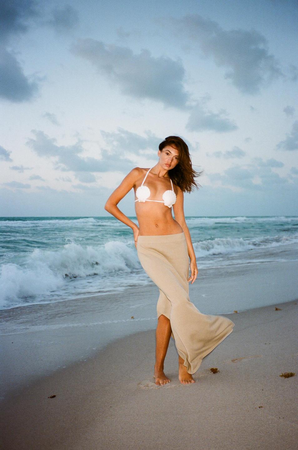 A woman wearing a seashell top and a long skirt stands on a beach with waves crashing behind her.