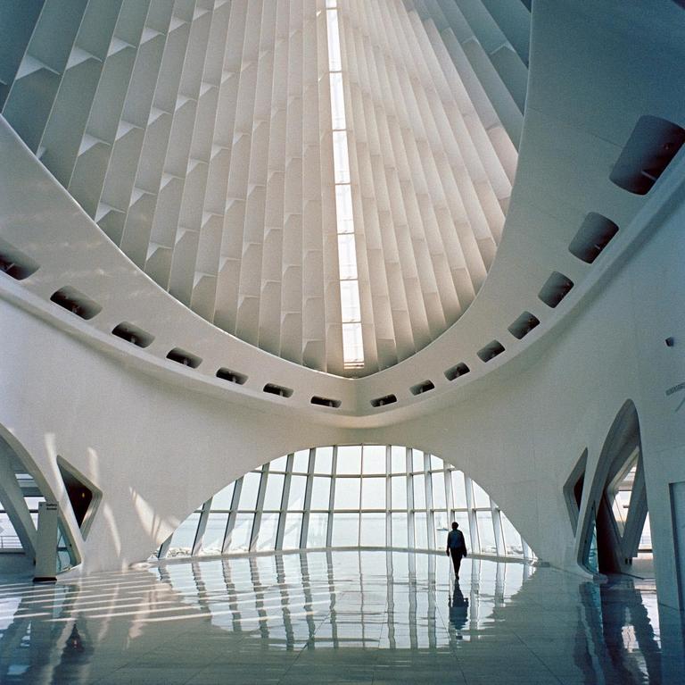A person walks across a reflective floor in a large, modern, white building with a high, textured ceiling and a curved glass wall overlooking water.