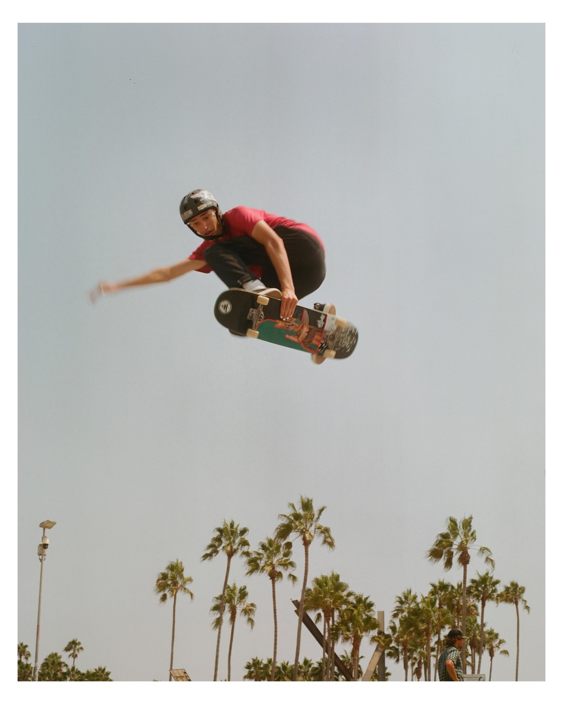 A man on a skateboard is airborne above palm trees and a street light.