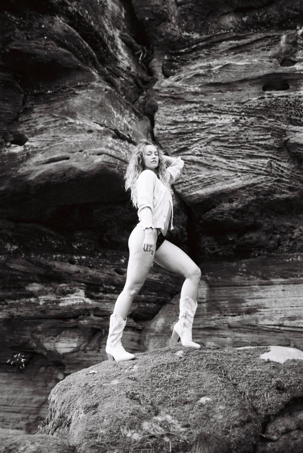 A woman in a cardigan and cowboy boots poses on a mossy rock in front of a large rock formation.