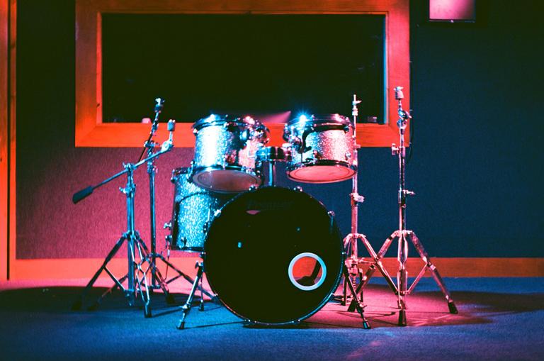 A drum kit with microphones stands in a room with a window, illuminated by blue and red light.