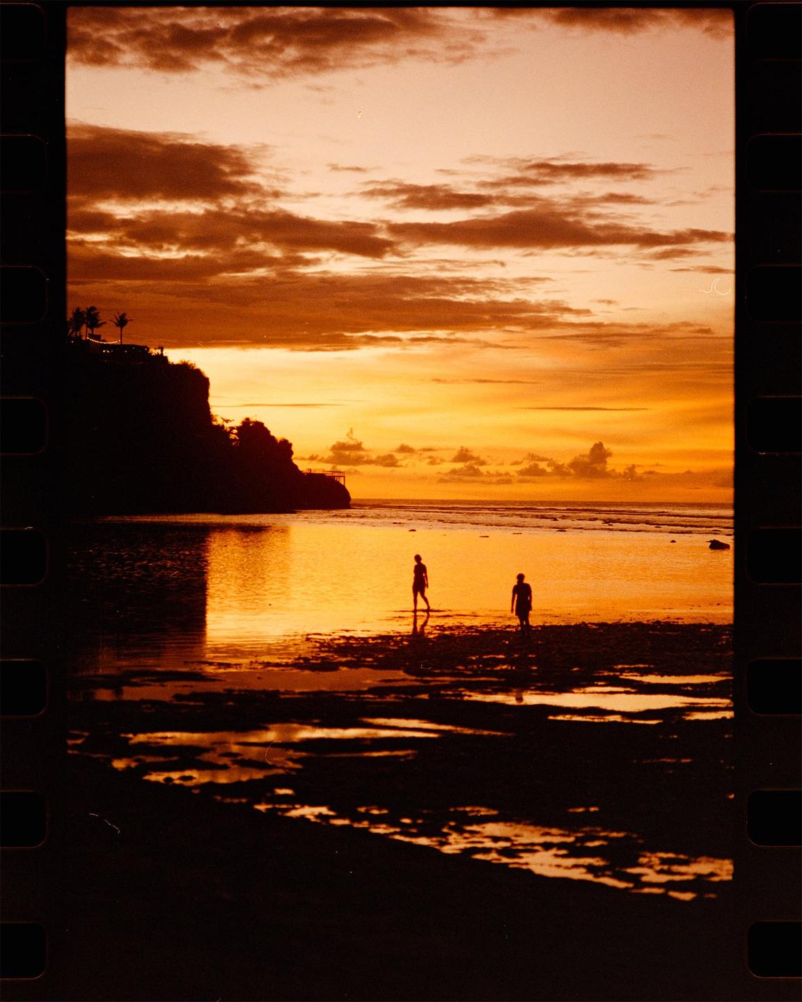 Two people walk in shallow water on a beach at sunset with a cliff and palm trees in the background.