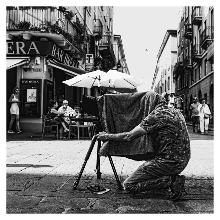 A man kneels on a cobblestone street, operating a large format camera covered with a dark cloth, with a cafe and people in the background.