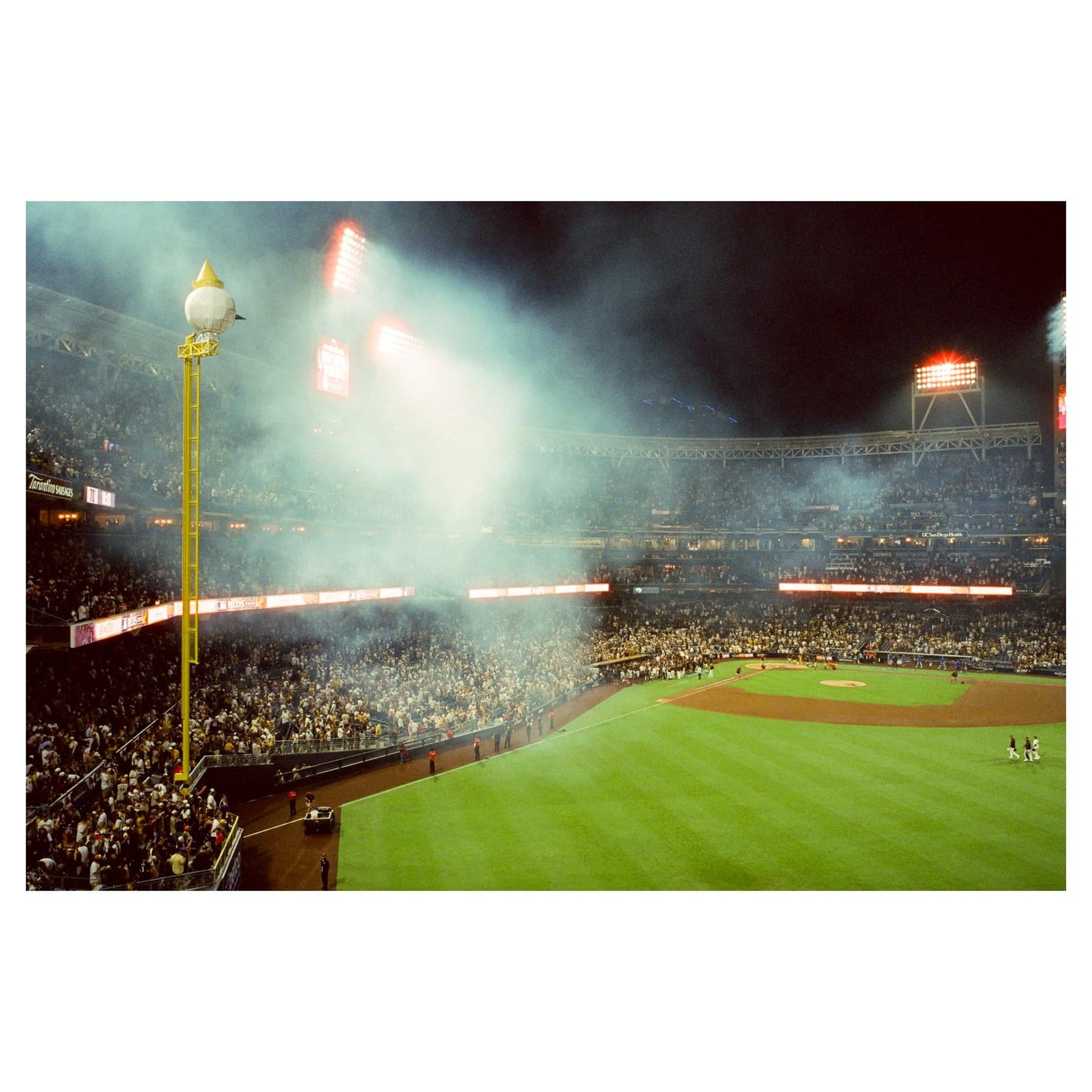 A baseball stadium is filled with spectators and smoke under bright lights at night.