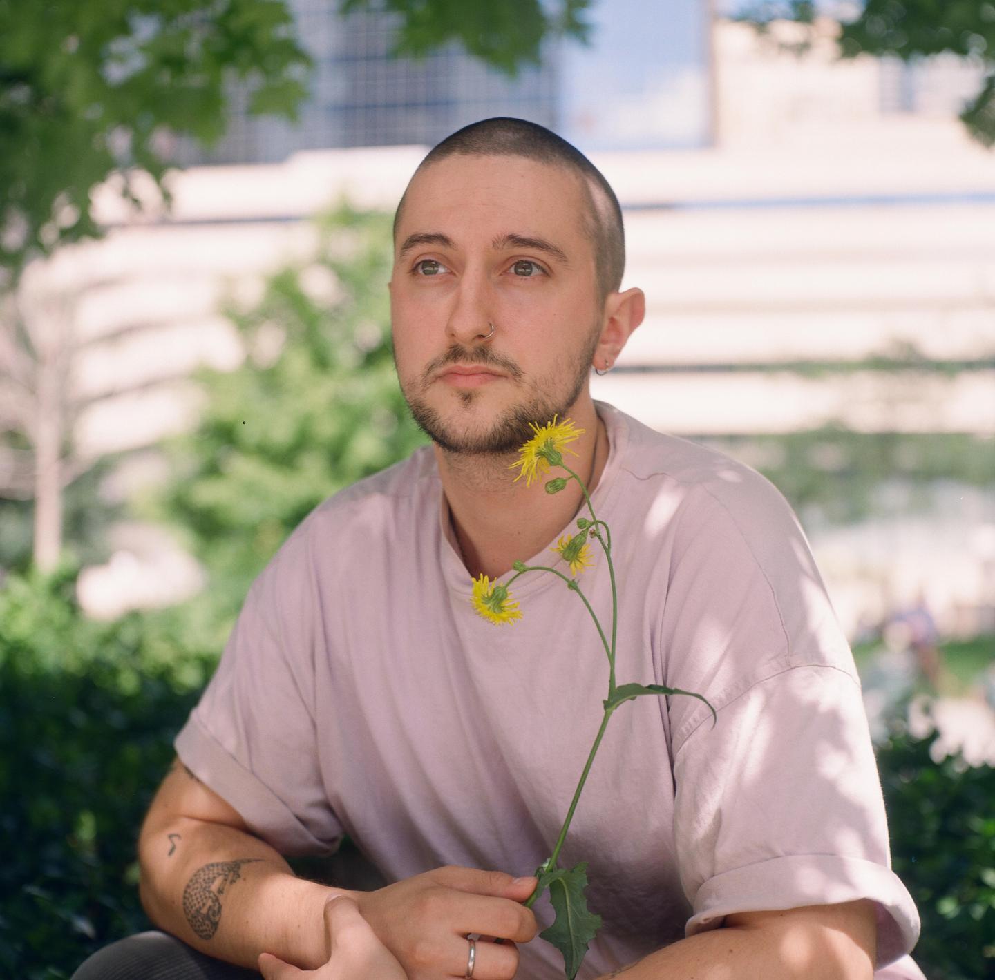 A man with a shaved head and beard holds a yellow flower and looks away from the camera.