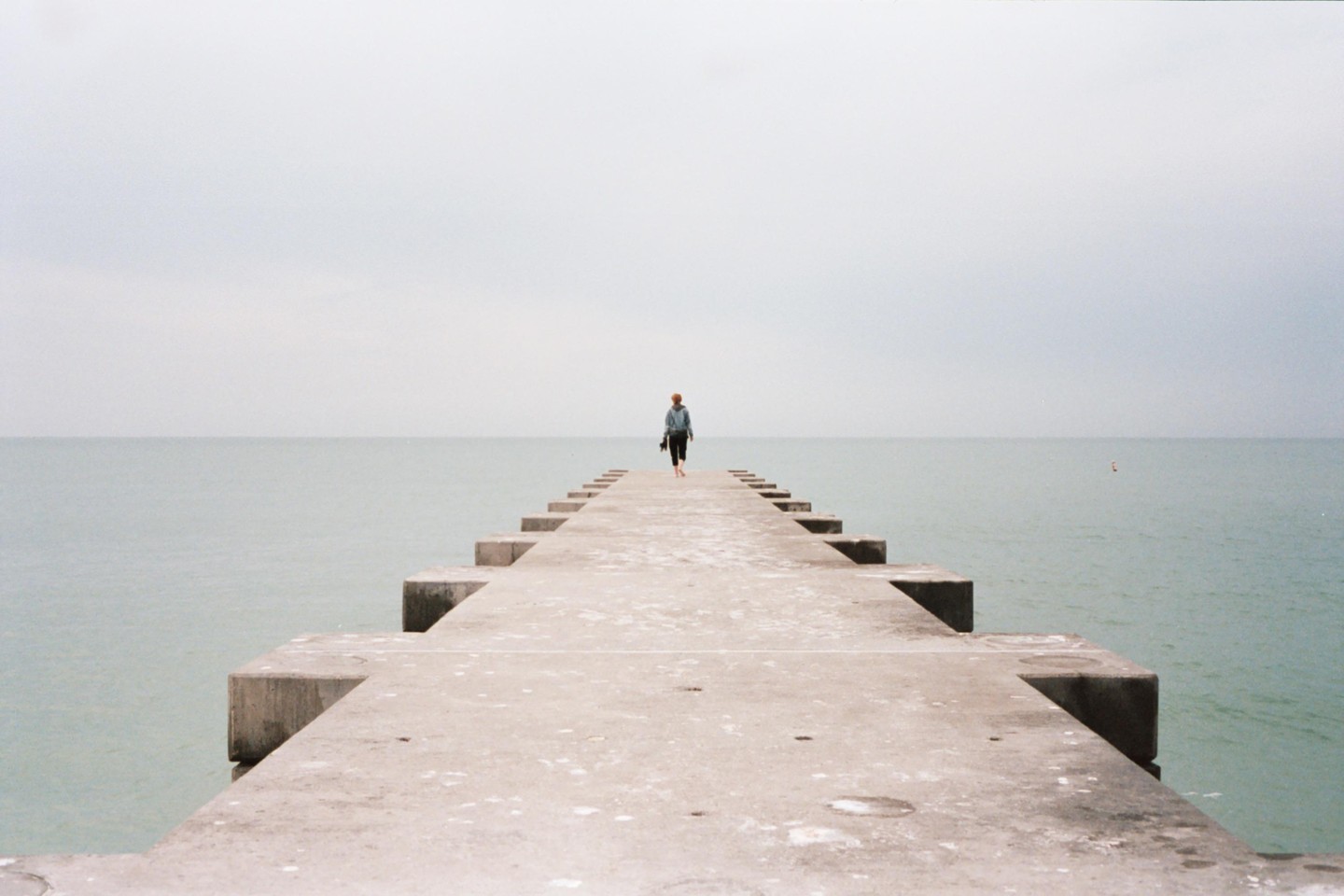 A person walks barefoot down a concrete pier extending into a calm body of water under an overcast sky.
