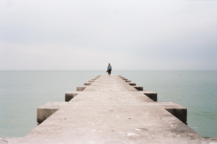 A person walks barefoot down a concrete pier extending into a calm body of water under an overcast sky.