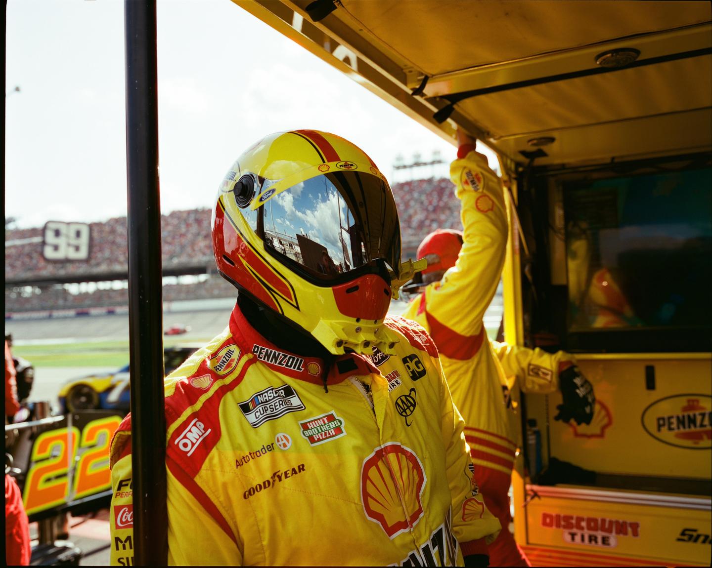 A man in a yellow racing suit and helmet stands in a pit stop area with a race track and spectators in the background.