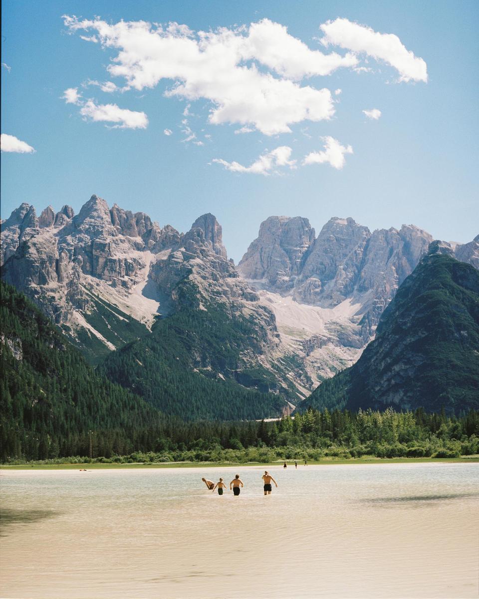 Several people wade in a shallow lake with a forest and mountains in the background under a blue sky with clouds.
