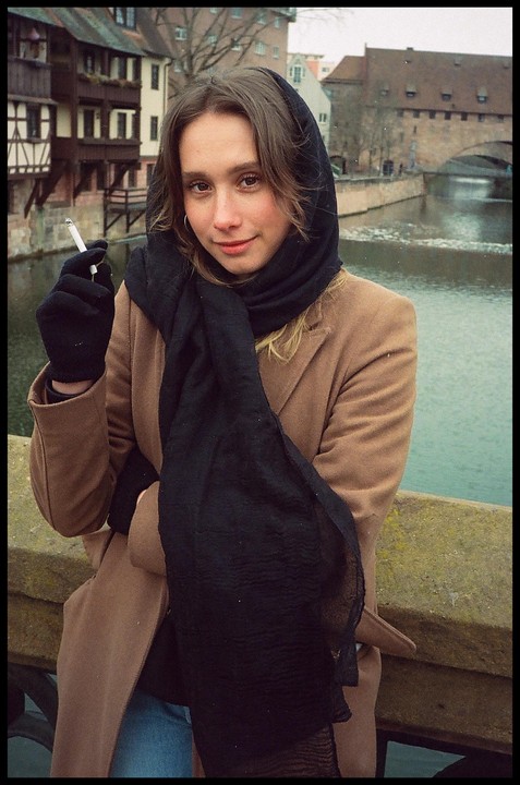 A woman wearing a black headscarf and a brown coat stands by a river, holding a cigarette.