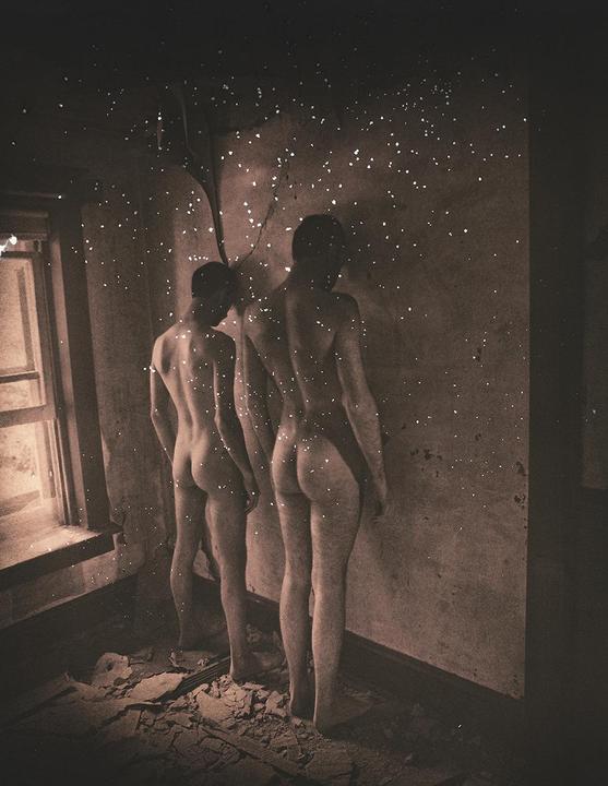 Two nude men stand in a derelict room with light particles floating around them.