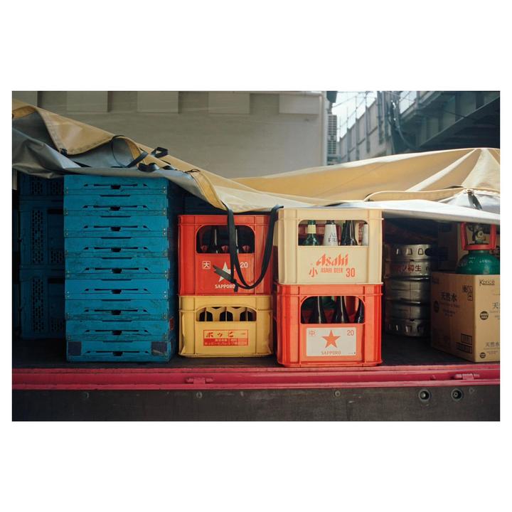 A truck bed is filled with stacks of blue crates, red and yellow beer crates, and other containers, covered by a tan tarp.