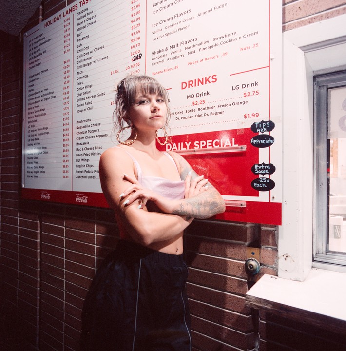 A woman with tattoos on her arms stands in front of a menu board with her arms crossed.