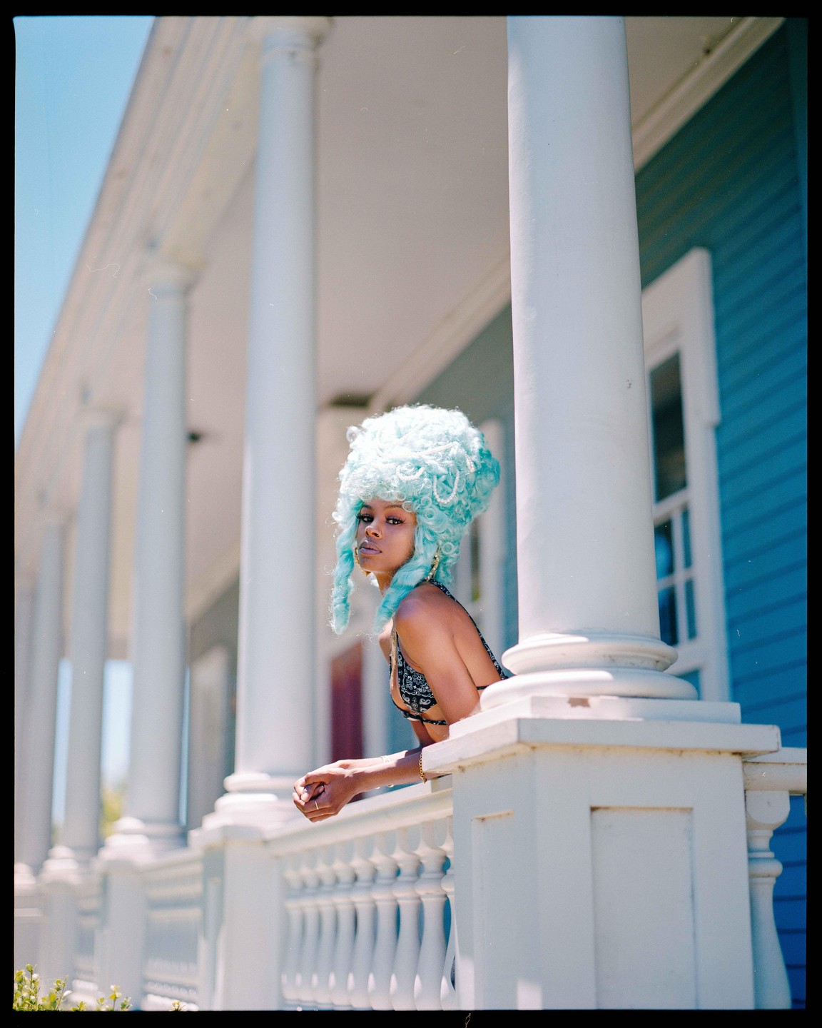 A woman with a large blue wig leans on a white railing on a porch with columns.