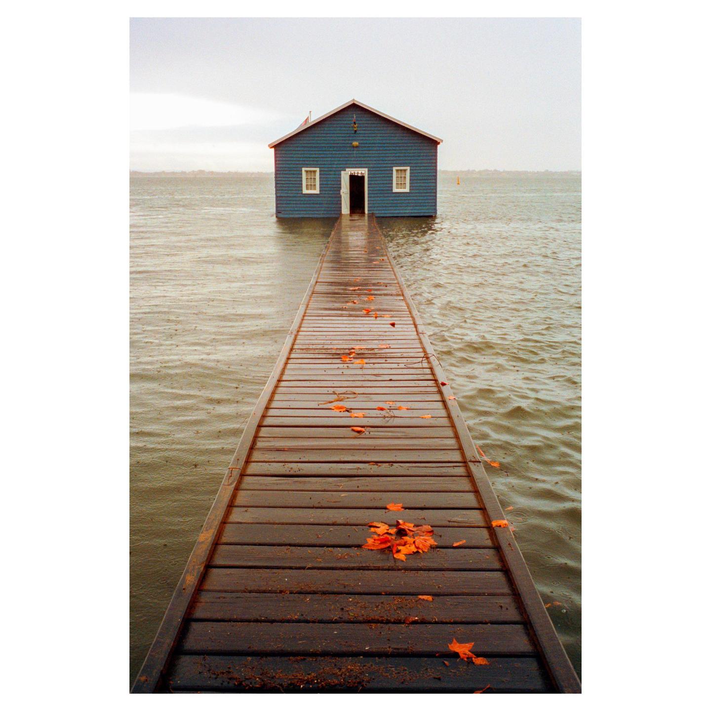 A long wooden jetty leads to a blue boathouse on the water under an overcast sky.