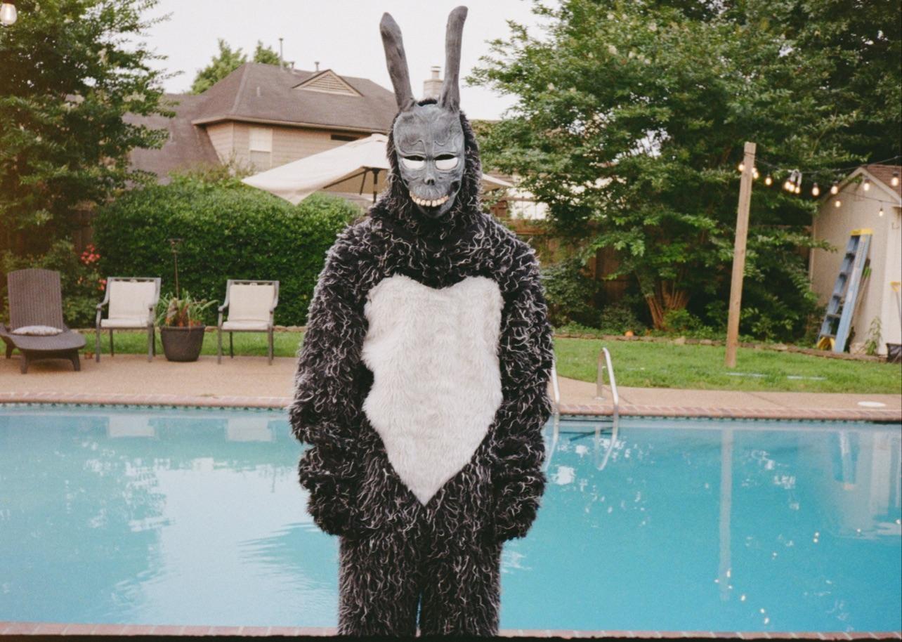 A person in a rabbit costume with a skull-like mask stands by a swimming pool in a backyard.