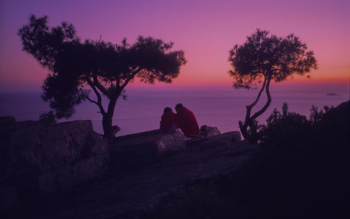 A couple sits on a rocky cliff overlooking the ocean at sunset, framed by two trees.