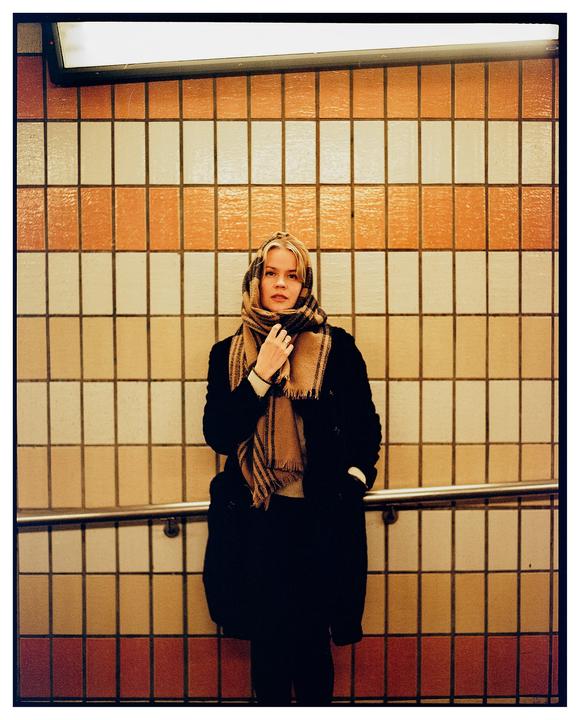 A woman wearing a plaid scarf and dark coat stands in front of a tiled wall.