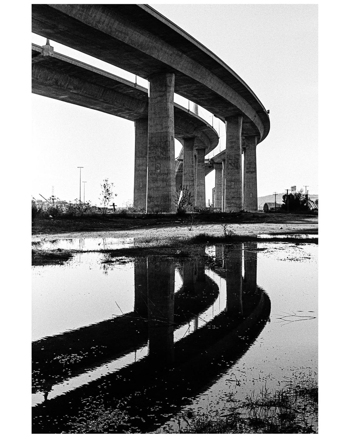 A concrete overpass with multiple levels is reflected in a large puddle on the ground.