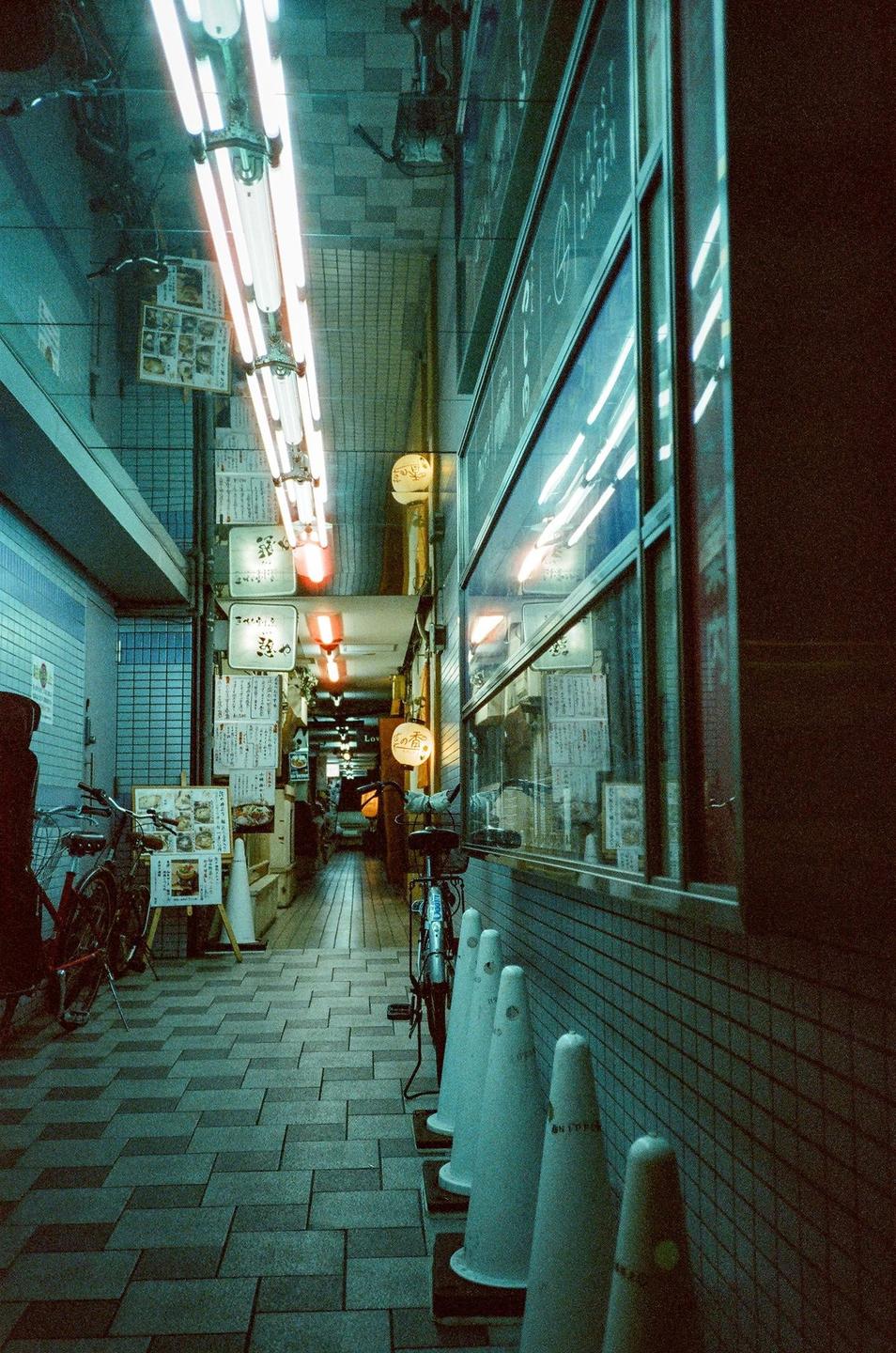 A narrow, neon-lit alleyway in Japan is lined with shops and bicycles.
