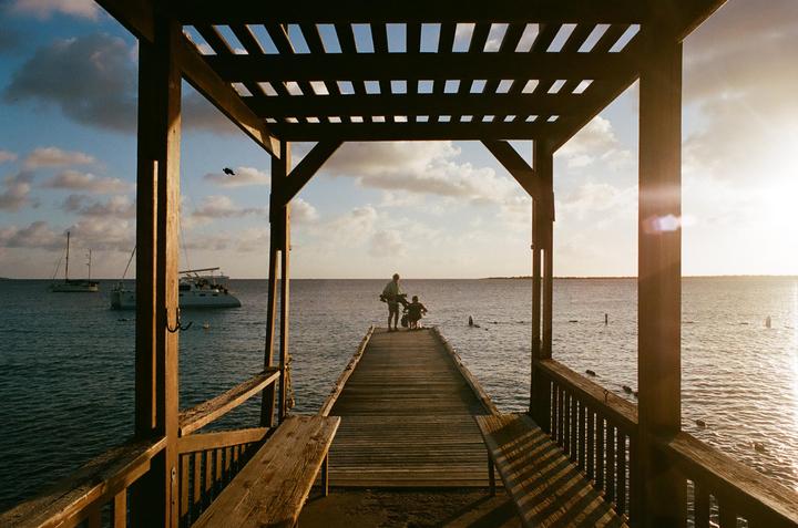 A man and a child stand on a wooden pier looking out at the ocean with boats in the distance under a cloudy sky at sunset.