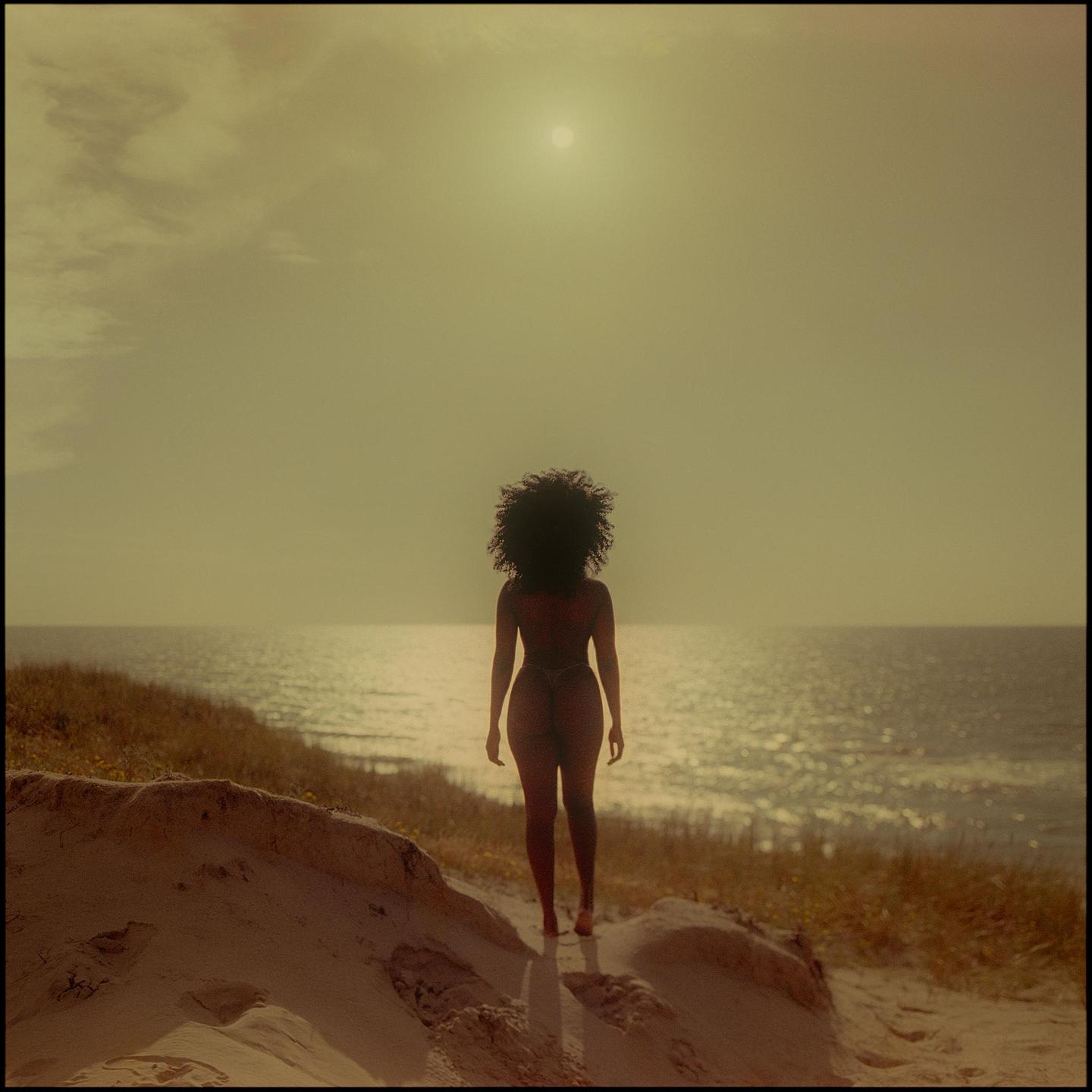 A woman with curly hair stands on a sandy dune looking out at the ocean under a hazy sun.