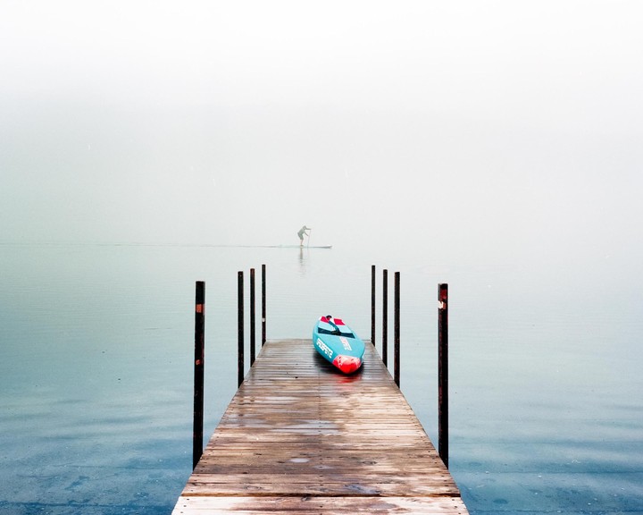 A blue paddleboard rests on a wooden dock while a person paddleboards in the distance on a hazy body of water.