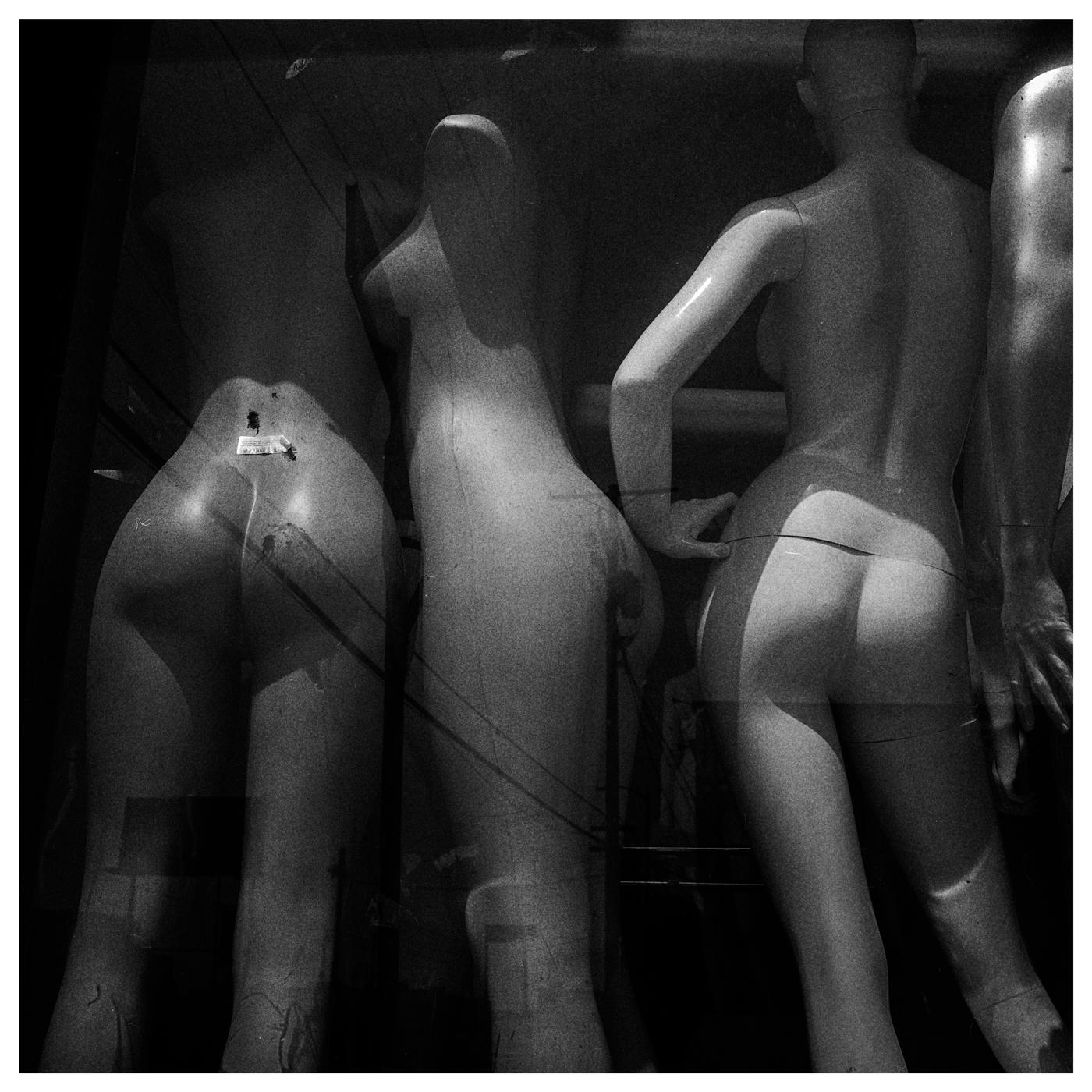 Three mannequins are visible from behind in a shop window, with reflections of power lines and buildings superimposed on them.