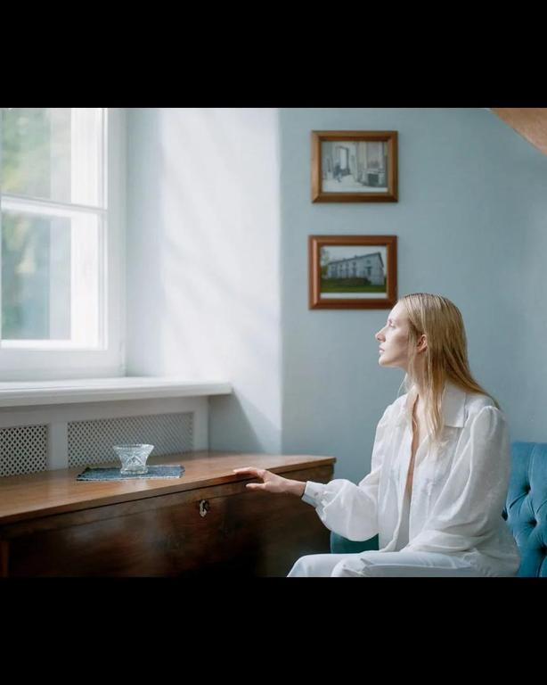 A woman in a white shirt sits on a blue sofa next to a wooden cabinet, looking out a window.