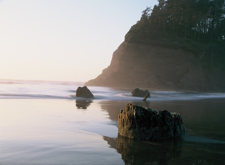 The ocean washes over a sandy beach with several rocks and a large cliff covered in trees.