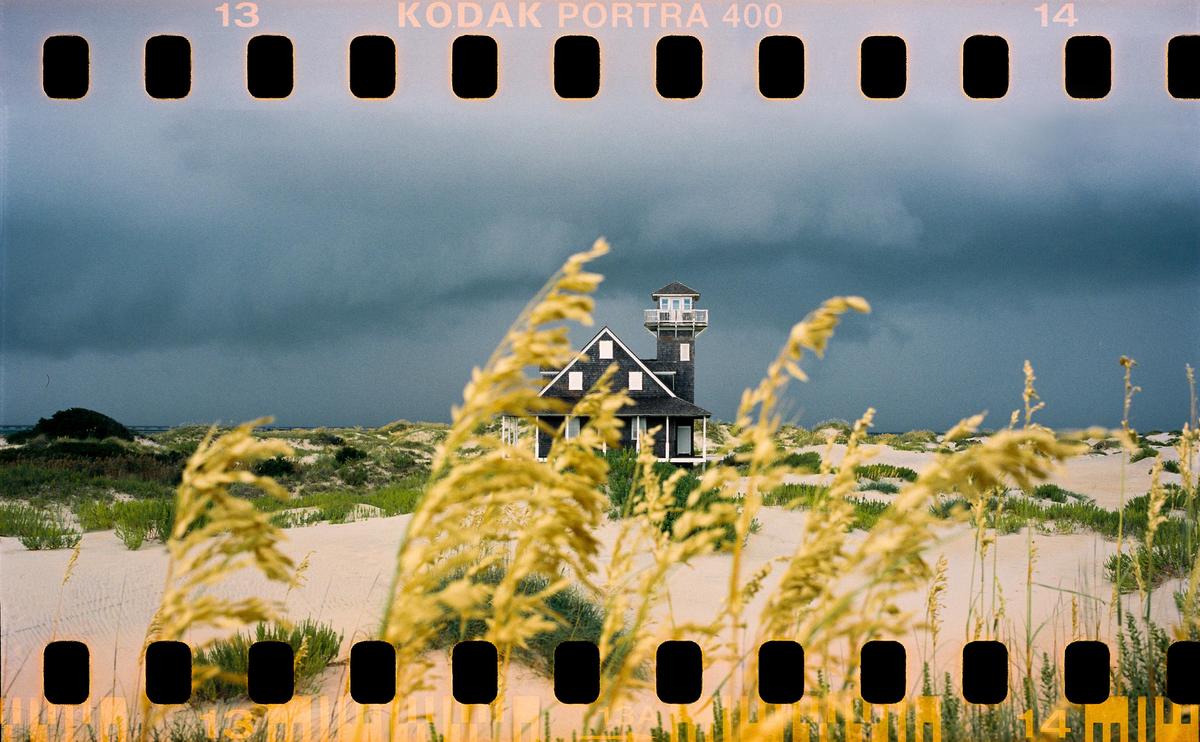 A house with a tower stands among sand dunes and tall grass under a stormy sky.