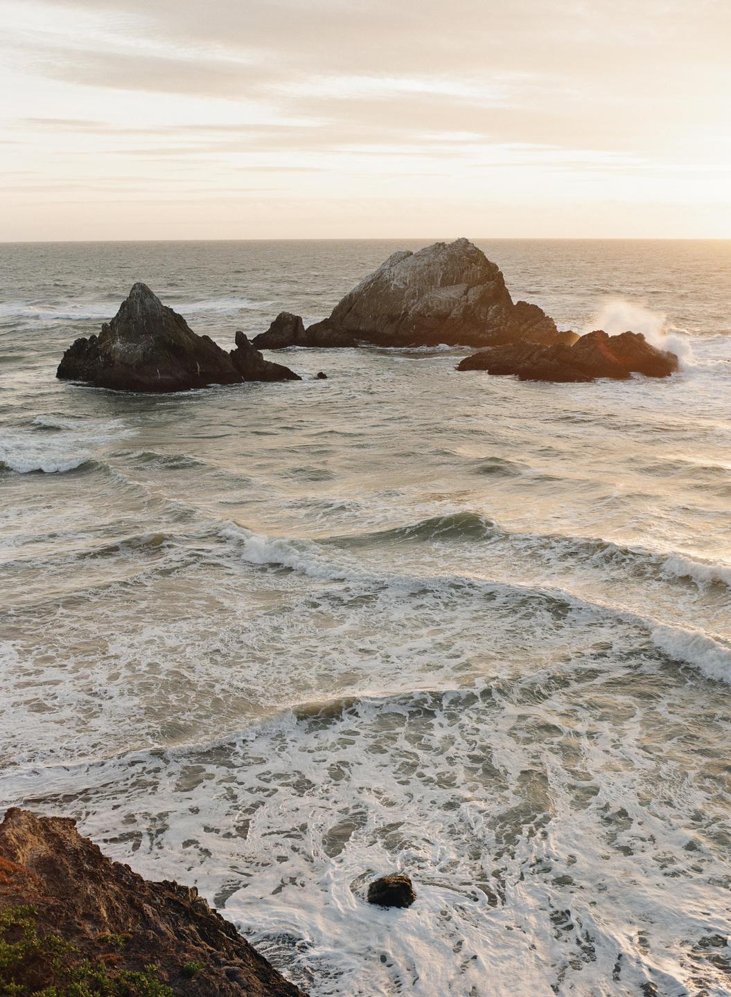 Waves crash against several large rocks in the ocean under a bright sky.