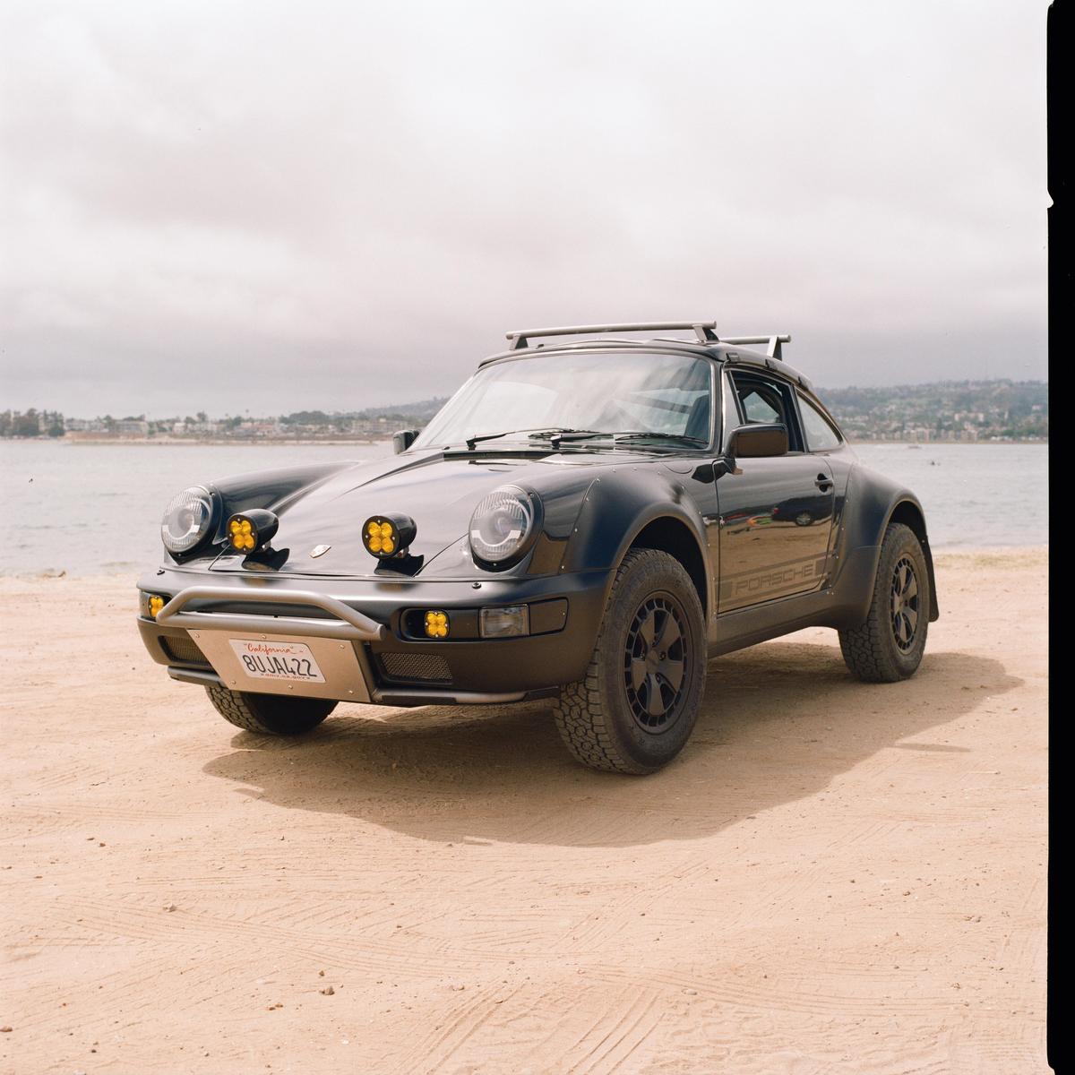 A black Porsche 911 with off-road modifications is parked on a sandy beach next to the ocean.
