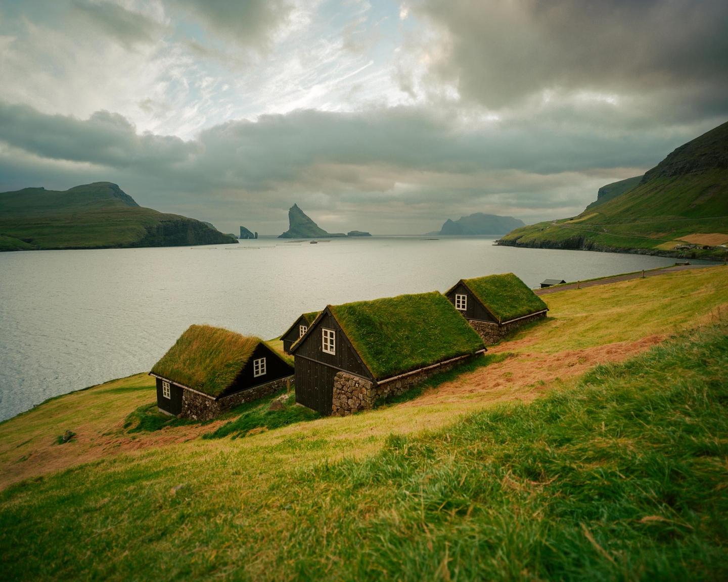 Three houses with grass roofs sit on a grassy hillside overlooking a body of water with mountains in the distance under a cloudy sky.