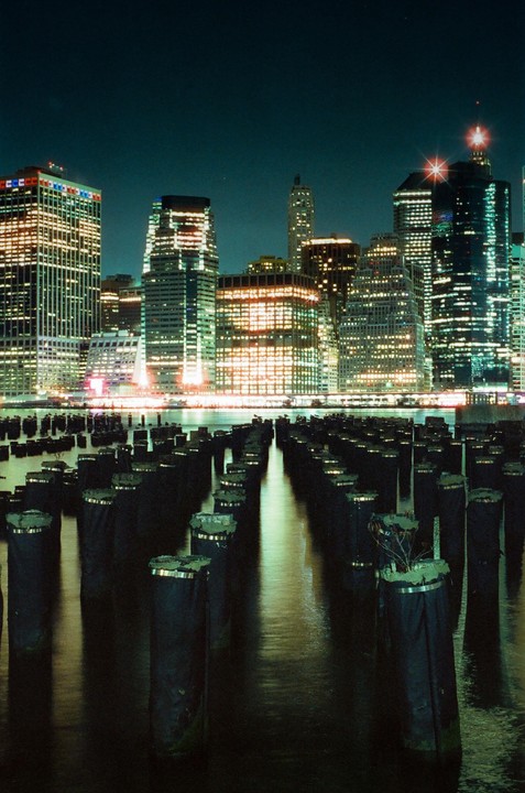 A long exposure captures the illuminated skyline of New York City at night, with rows of dark pier pilings in the foreground.