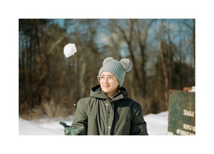 A woman in a winter coat and hat throws a snowball in a snowy forest.