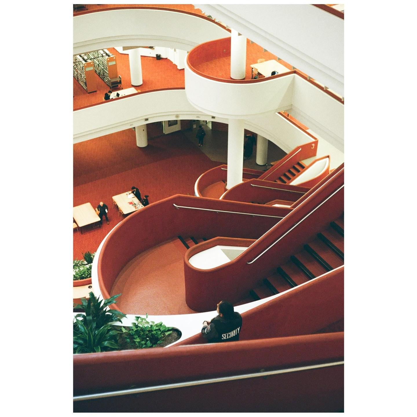 A person wearing a "SECURITY" jacket sits on a red staircase in a large, multi-level building with red carpet and white railings.