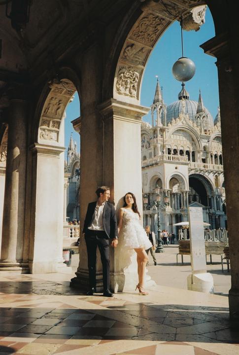 A couple stands under an archway with a large ornate building in the background.