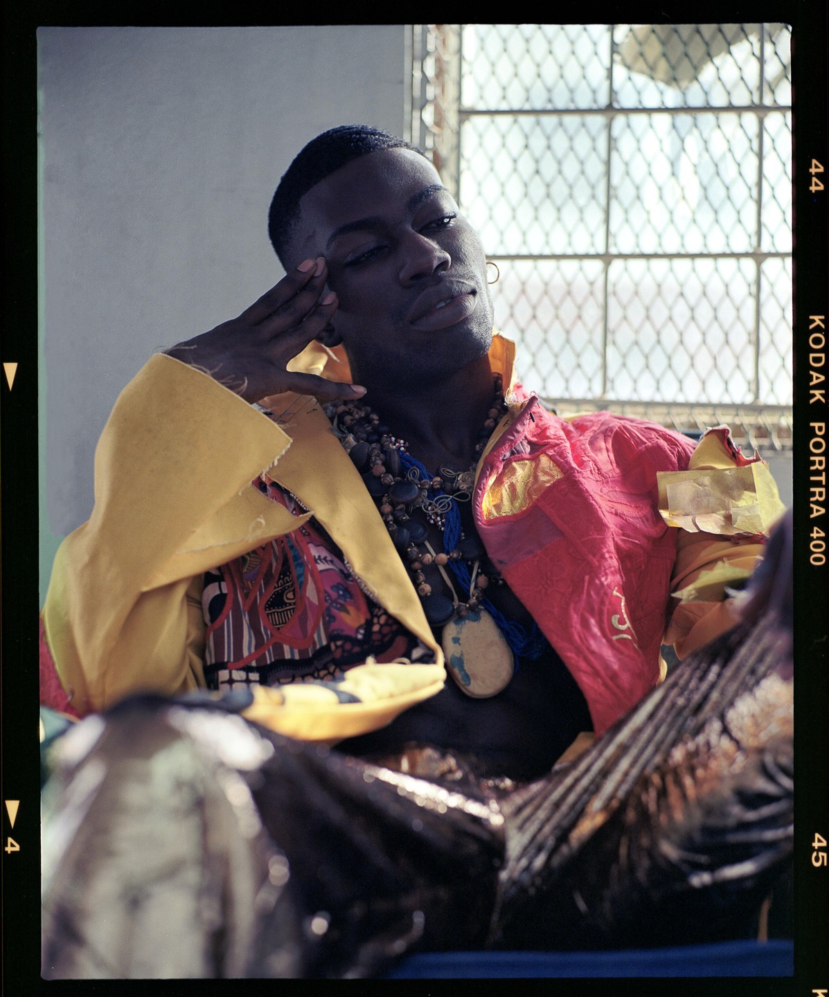 A man wearing colorful clothing and many necklaces poses in front of a window with a metal grate.