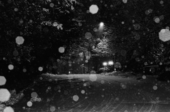 A snowy street at night is illuminated by streetlights, with large snowflakes visible in the foreground.