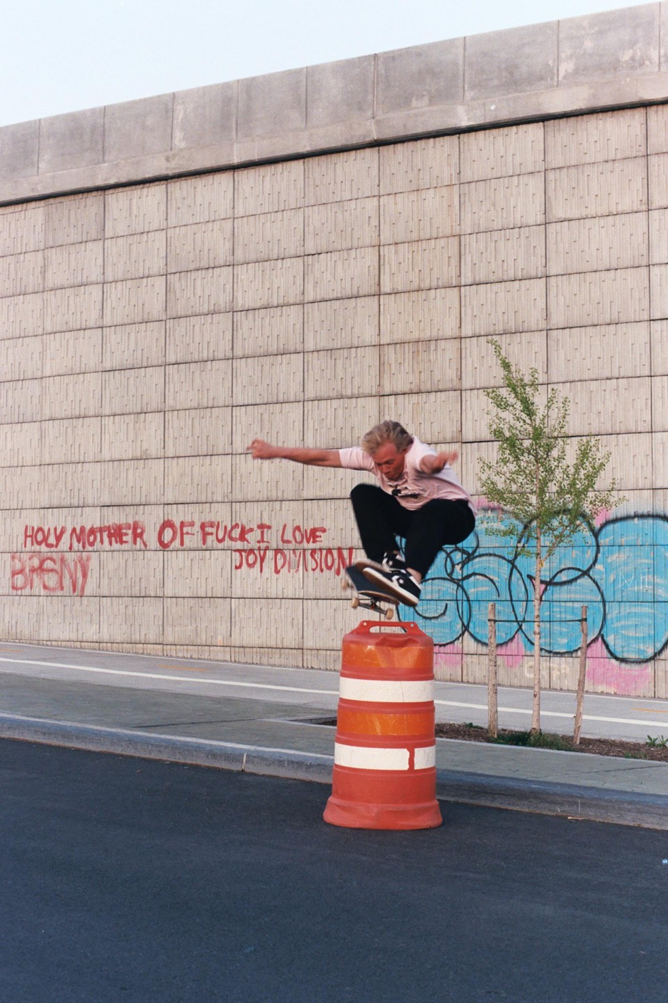 A man on a skateboard jumps over an orange barrel in front of a concrete wall with graffiti.