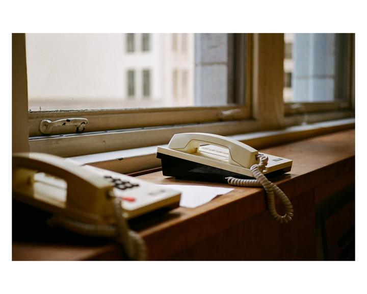 Two landline telephones sit on a wooden desk in front of a window looking out at a building.