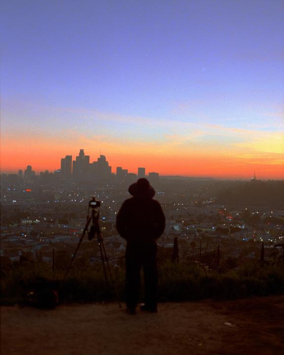 A person stands on a hill overlooking the Los Angeles skyline at sunset, with a tripod set up nearby.