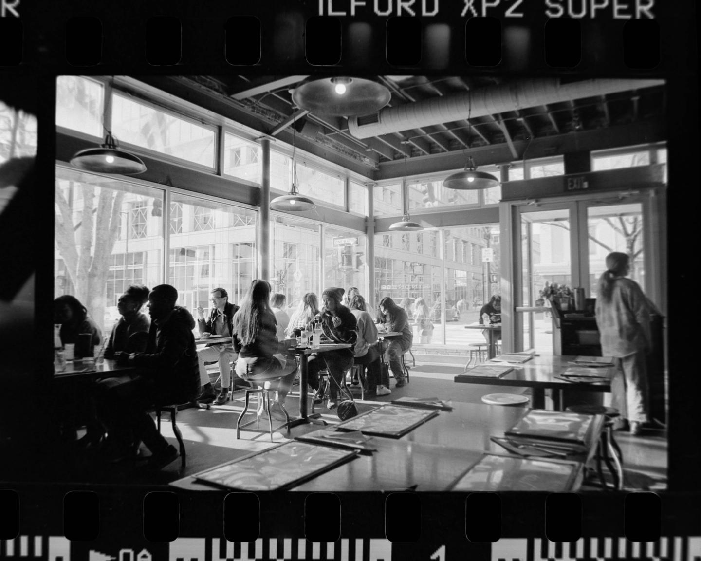 People sit at tables in a brightly lit cafe with large windows looking out onto a street.